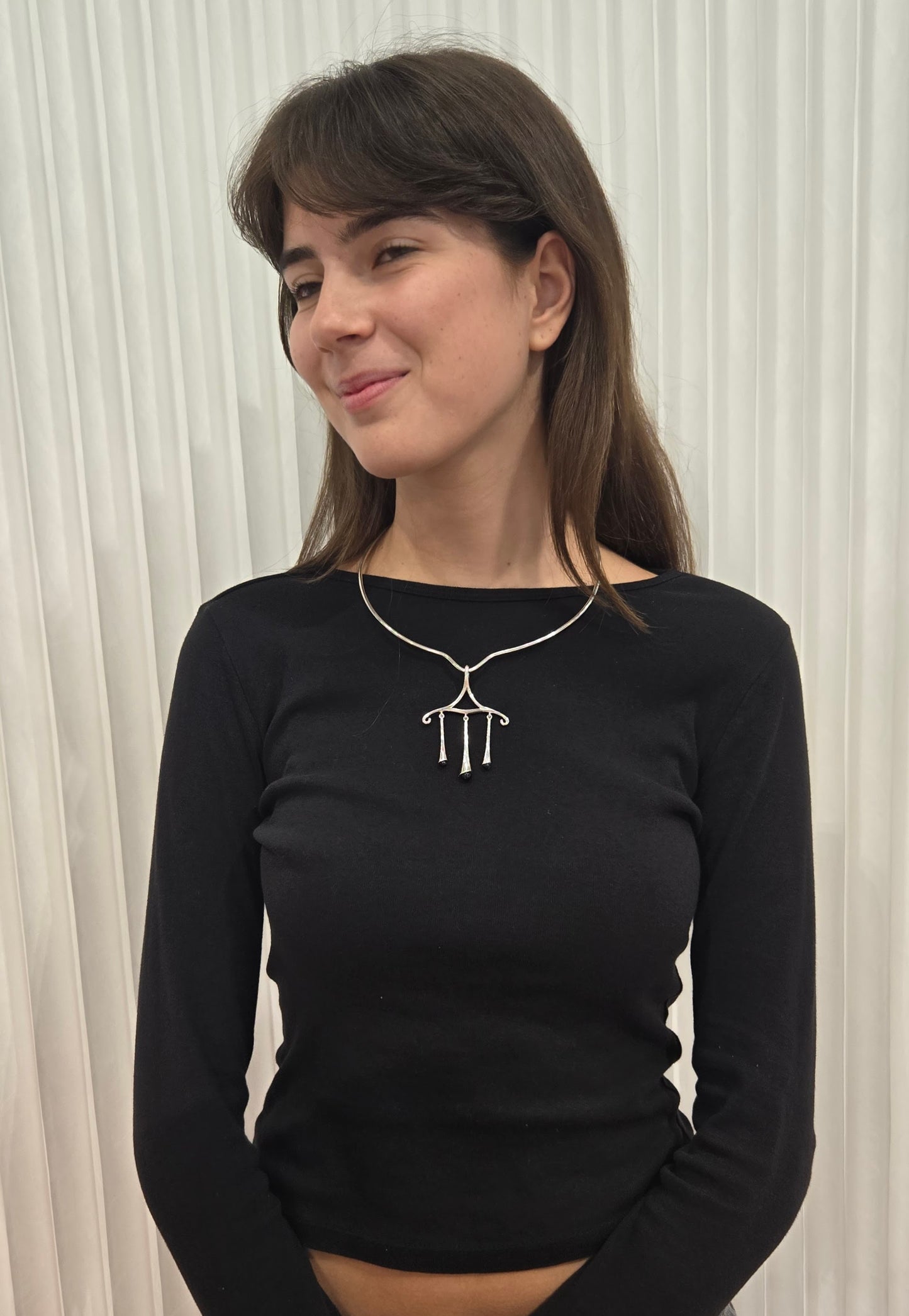 Woman wearing a black long-sleeve top with chandelier pendant necklace like sophie buhai against a white curtain background
