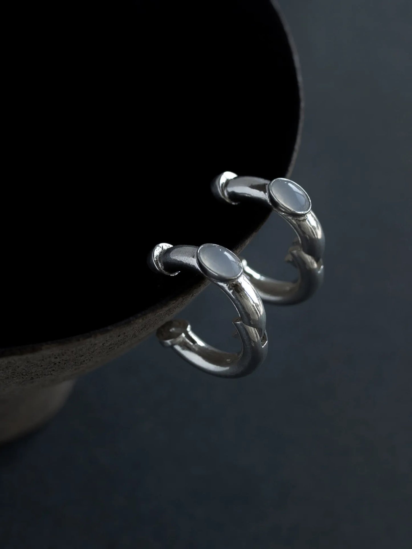 Maancy modernist silver hoop earrings with interchangeable chalcedony cabochons displayed on dark surface, showing sculptural design.