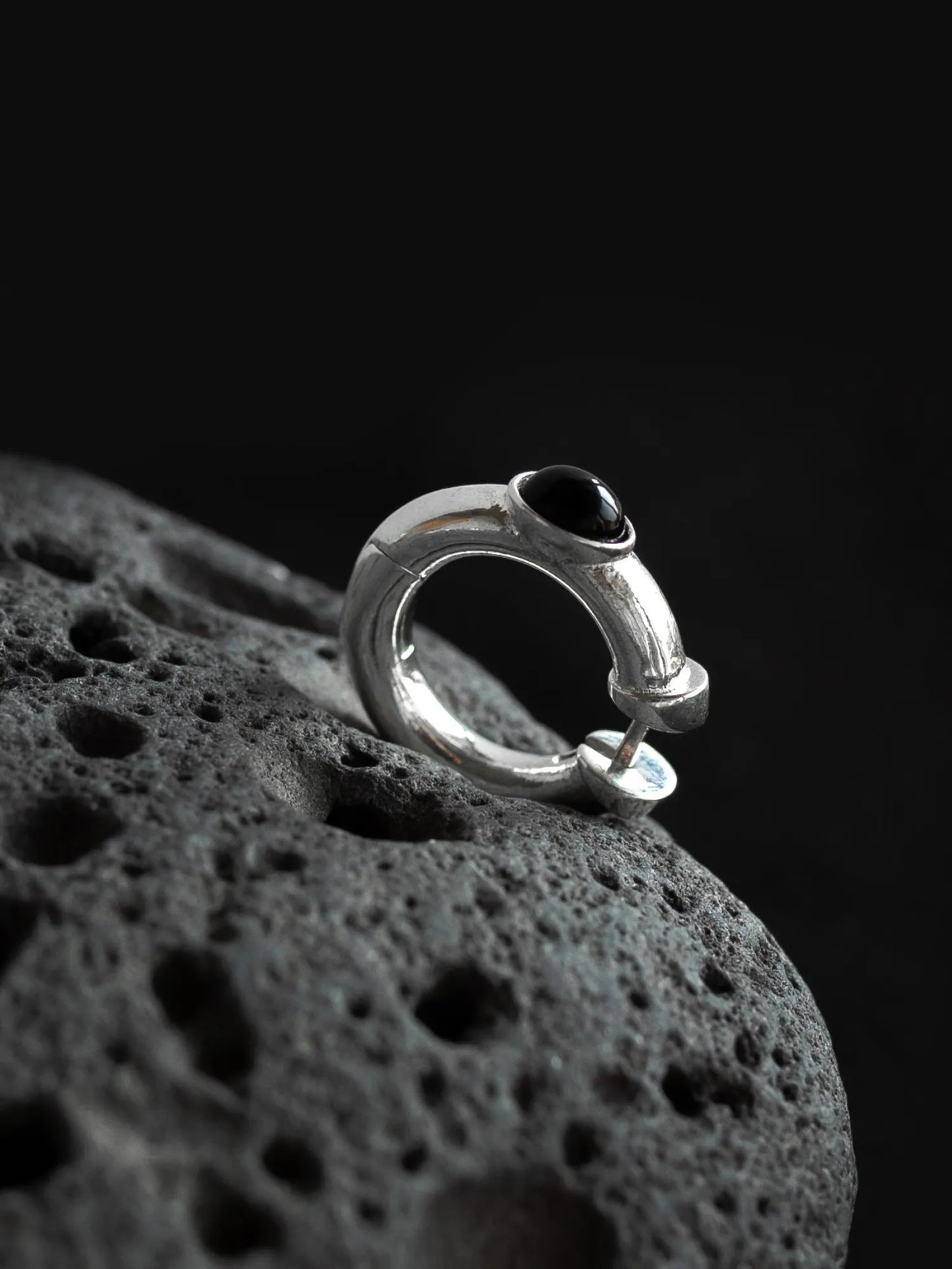 Close-up detail of Maancy modernist silver hoop earring with onyx stone on volcanic texture, highlighting architectural jewelry design.