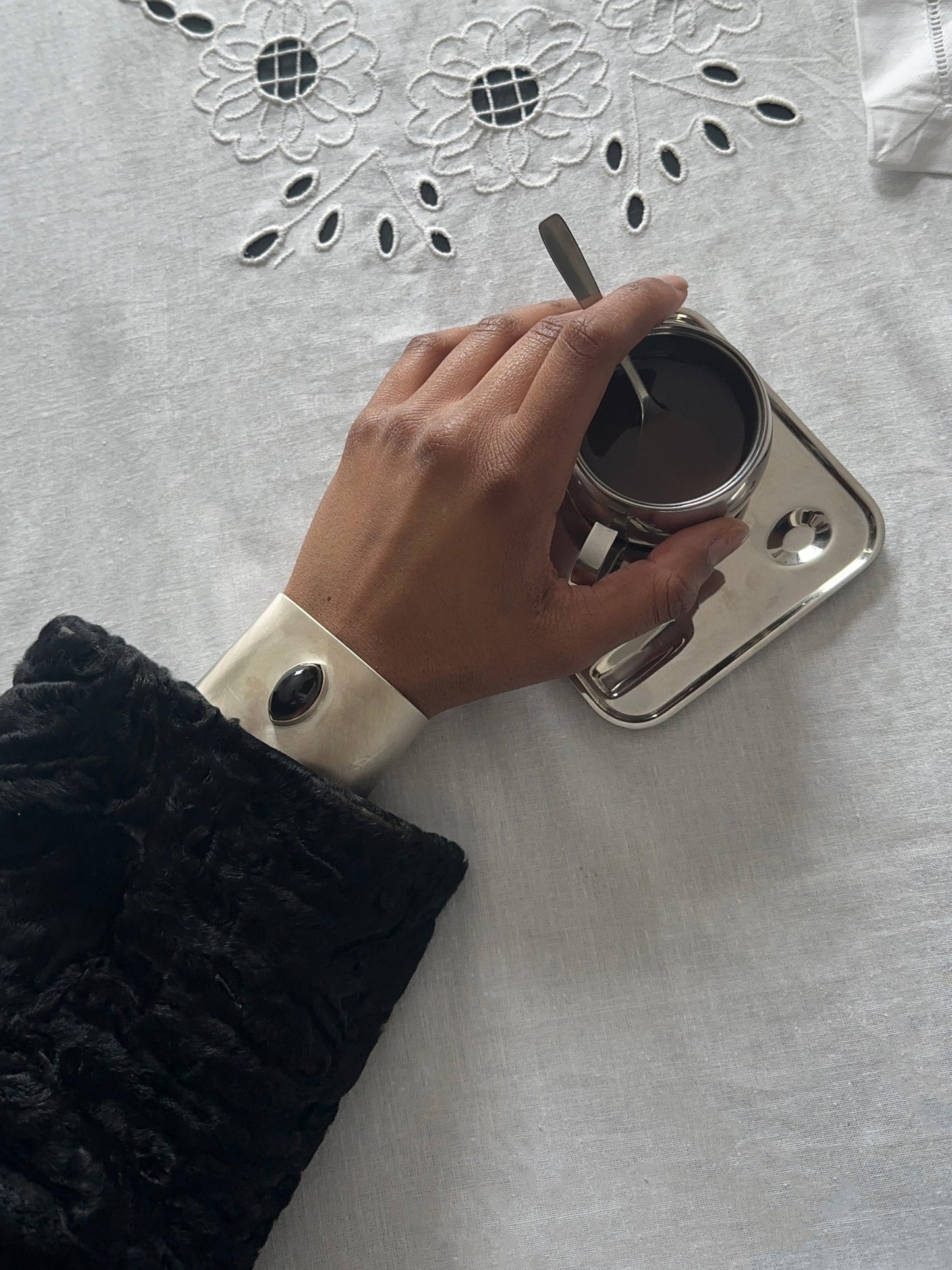 Hand holding a silver cup with a dark beverage on a textured surface wearing marquise cuff bangle, architectural heirloom