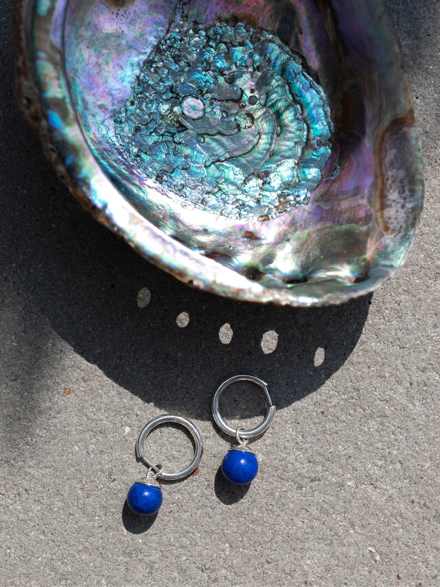 Maancy sterling silver hoop earrings with lapis drops displayed with iridescent abalone shell in natural light, emphasizing color harmony and artisan craftsmanship.